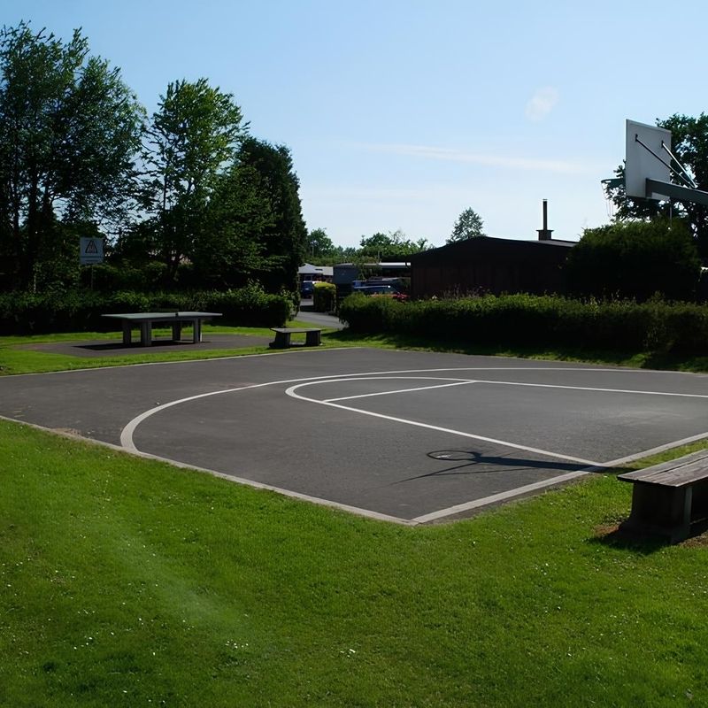 Ein Basketballplatz mit einem Basketballkorb und im Hintergrund ist eine Tischtennisplatte zu sehen.